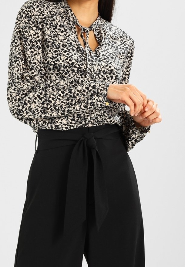 Femme portant un chemisier floral noir et blanc avec un col à nouer et un pantalon noir taille haute avec une ceinture à nœud à l'avant.