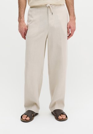 Mann trägt beige weite Hosen mit Kordelzug in der Taille und schwarze Slide-Sandalen, steht vor weißem Hintergrund.