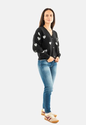 Femme debout les mains jointes, portant un cardigan noir avec des cœurs blancs, un jean bleu et des baskets blanches avec des rayures brunes.