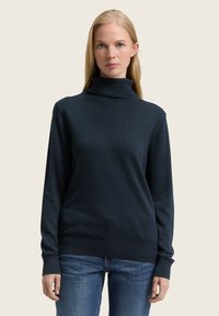 Pull à col roulé marine en tricot doux, avec des manches longues, des poignets côtelés et une silhouette ajustée. Porté avec un jean bleu.