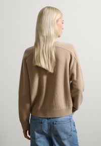 Beige gebreide trui met een relaxte fit, verlaagde schouders en geribbelde zoom. Gecombineerd met lichtblauwe high-waisted jeans, wat een casual stijl laat zien.