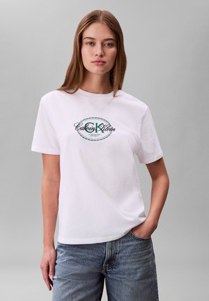 Jeune femme portant un t-shirt blanc Calvin Klein avec un imprimé logo vert, jean bleu, debout devant un fond gris uni.