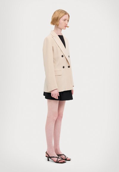 Blazer beige à double boutonnage avec des boutons noirs, texture lisse, associé à une mini-jupe noire et des sandales à talons noirs à lanières.
