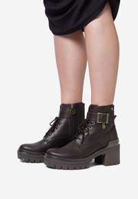 Botine din piele maro, cu toc gros, detalii cu șireturi, fermoar lateral și accent cu cataramă. Talpă texturată pentru aderență și durabilitate.