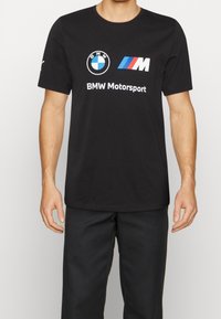 Muž oblečený v černém tričku BMW Motorsport s logem BMW a M pruhy, doplněný černými kalhotami na jednoduchém pozadí.