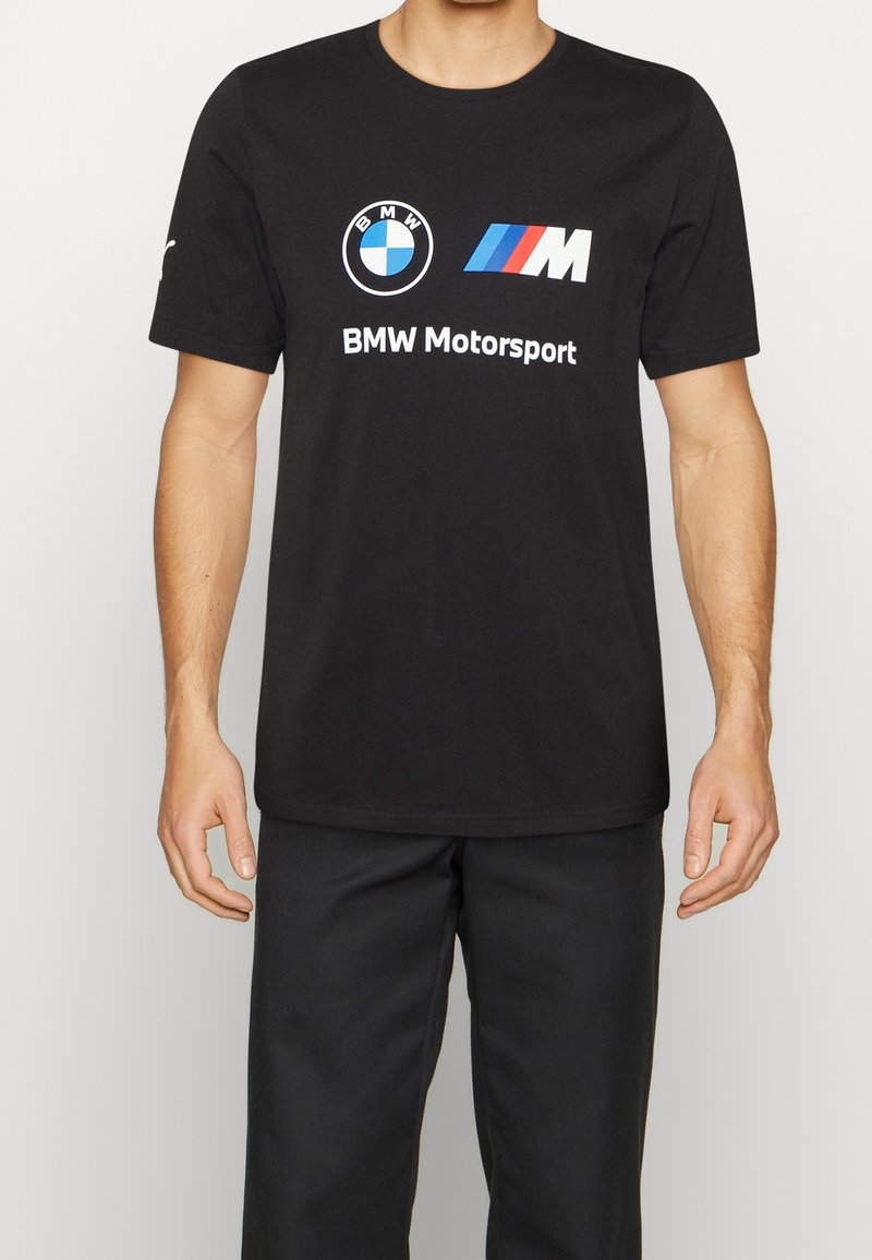 Muž oblečený v černém tričku BMW Motorsport s logem BMW a M pruhy, doplněný černými kalhotami na jednoduchém pozadí.