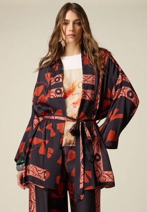 Donna che indossa una giacca larga nera e rossa a fantasia floreale e pantaloni abbinati, con una camicia beige stampata sotto e braccialetti verdi.