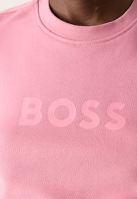 Ružová mikina s okrúhlym výstrihom, so širokým, embosovaným logom "BOSS" v zhodnej farbe cez hrudník. Mäkká textúra látky.