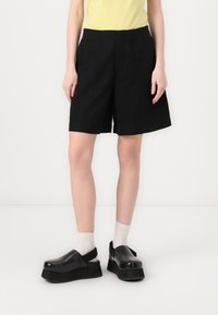 Shorts noirs en tissu léger, avec une coupe droite et des plis. Associés à des sabots noirs épais et des chaussettes blanches à la cheville.