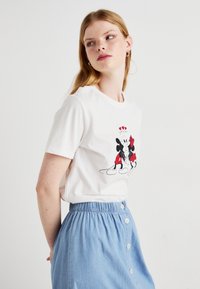 T-shirt bianco in cotone con una grafica di due topolini dei cartoni animati con dettagli rossi, abbinato a una gonna azzurro chiaro con bottoni.