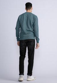 Tealfarbenes Langarm-Sweatshirt aus Baumwollmaterial, lockere Passform, gerippte Bündchen und Saum, kombiniert mit schwarzen Jeans und weißen Turnschuhen. Sichtbare Designdetails auf der Rückseite.