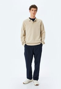 Vaale Beige -värinen pitkähihainen polo kauluksella, yhdistettynä tummansinisiin kapeneviin housuihin ja valkoisiin lenkkareihin. Asussa on rento istuvuus ja pehmeä tekstuuri.