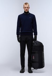 Zwarte rugzak met een opvallend merkembleem en een detail van de Noorse vlag. Draagt door een model in een marineblauwe trui, zwarte broek en leren handschoenen.