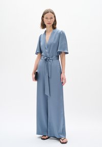 Hellblauer Jumpsuit mit Wickeldesign, kurzen Ärmeln, einem Bindegürtel in der Taille und weitem Bein. Glattes Textilgewebe.