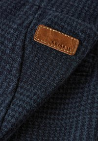 Marinblauer Textilstoff mit einem Hahnentrittmuster, versehen mit einem braunen Lederetikett, auf dem "name it" genäht ist. Sichtbare Nähte auf dem Etikett.