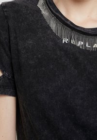 Camiseta negra de manga corta, con un acento de cristal brillante a lo largo del escote y una textura desvaída. El nombre de la marca es visible.