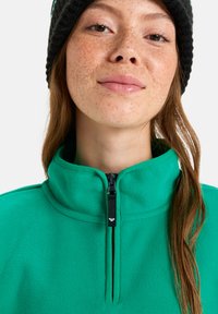 Pullover de polar verde com gola alta e fecho éclair preto. Apresenta uma textura suave e detalhes de costura discretos. Completo com um gorro preto.