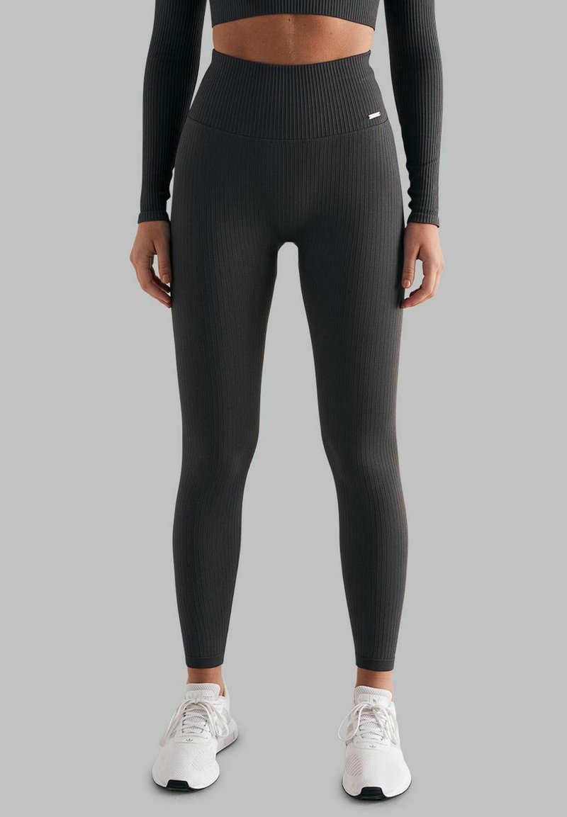 Ribbe svarte tights med høy midje og ankellang design, parret med hvite joggesko på en grå bakgrunn.
