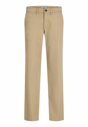 Ung dreng står iført en mørkeblå skjorte med knapper over en hvid T-shirt, beige bukser og hvide sneakers mod en ensfarvet lys baggrund.