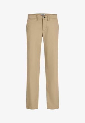Jeune garçon debout, portant une chemise bleu marine boutonnée sur un t-shirt blanc, un pantalon beige et des baskets blanches, sur un fond clair uni.