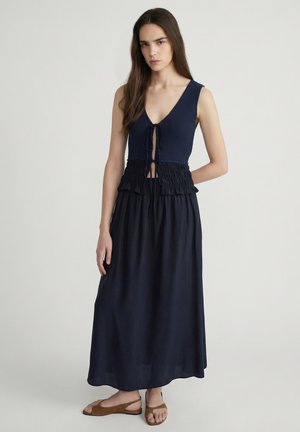 Femme aux longs cheveux bruns portant un haut sans manches bleu marine avec des détails noués et une longue jupe bleu marine, debout avec une expression neutre et des sandales marron.