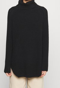 Person in einem schwarzen gestrickten Rollkragenpullover mit Waffelstruktur und beigen Hosen, die Hände entspannt an den Seiten.