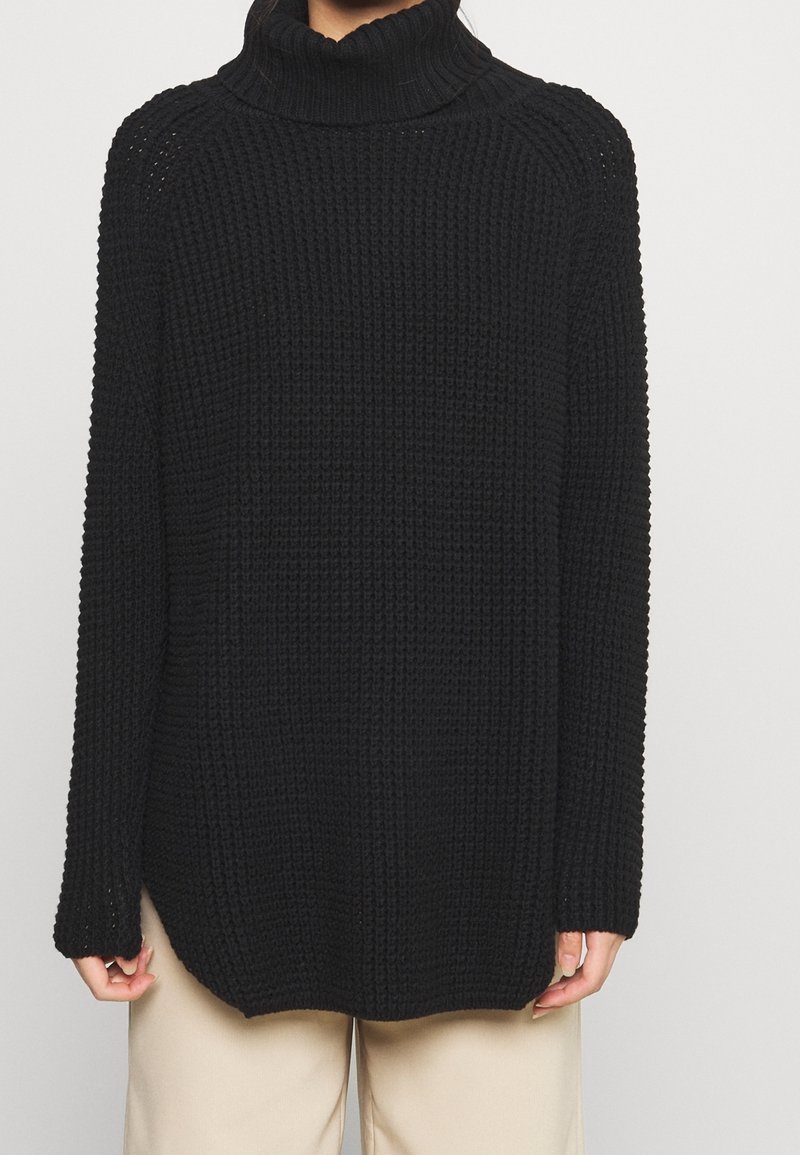 Person in einem schwarzen gestrickten Rollkragenpullover mit Waffelstruktur und beigen Hosen, die Hände entspannt an den Seiten.