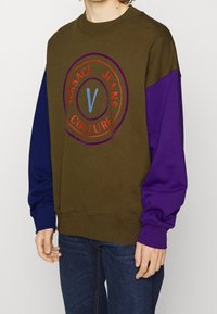 Olivgrön sweatshirt med lila ärmar som har en cirkulär logo i röd och blå text. Ribbad rundad halsringning och muddar.