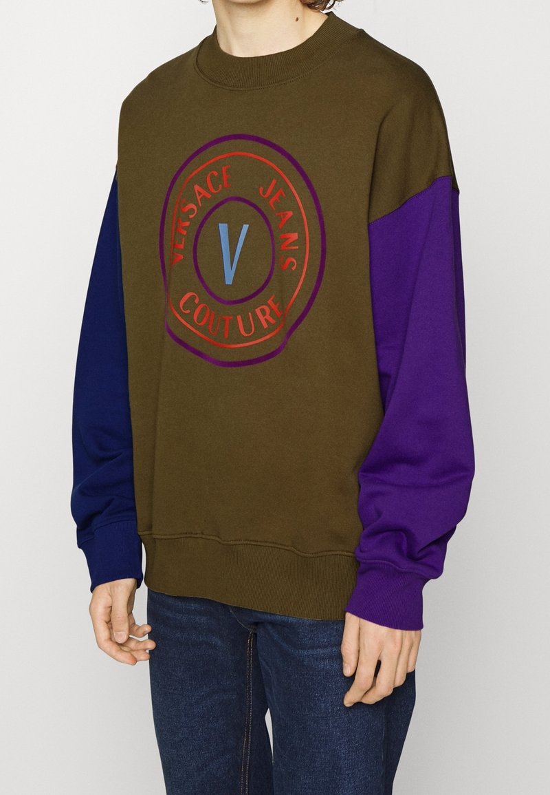 Olivgrön sweatshirt med lila ärmar som har en cirkulär logo i röd och blå text. Ribbad rundad halsringning och muddar.