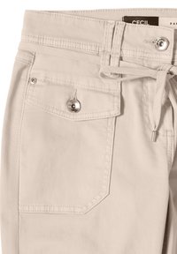 Pantalon beige avec une poche frontale à rabat fermée par un bouton argenté, détail de rivet argenté, passants de ceinture et un cordon de serrage assorti à la taille.