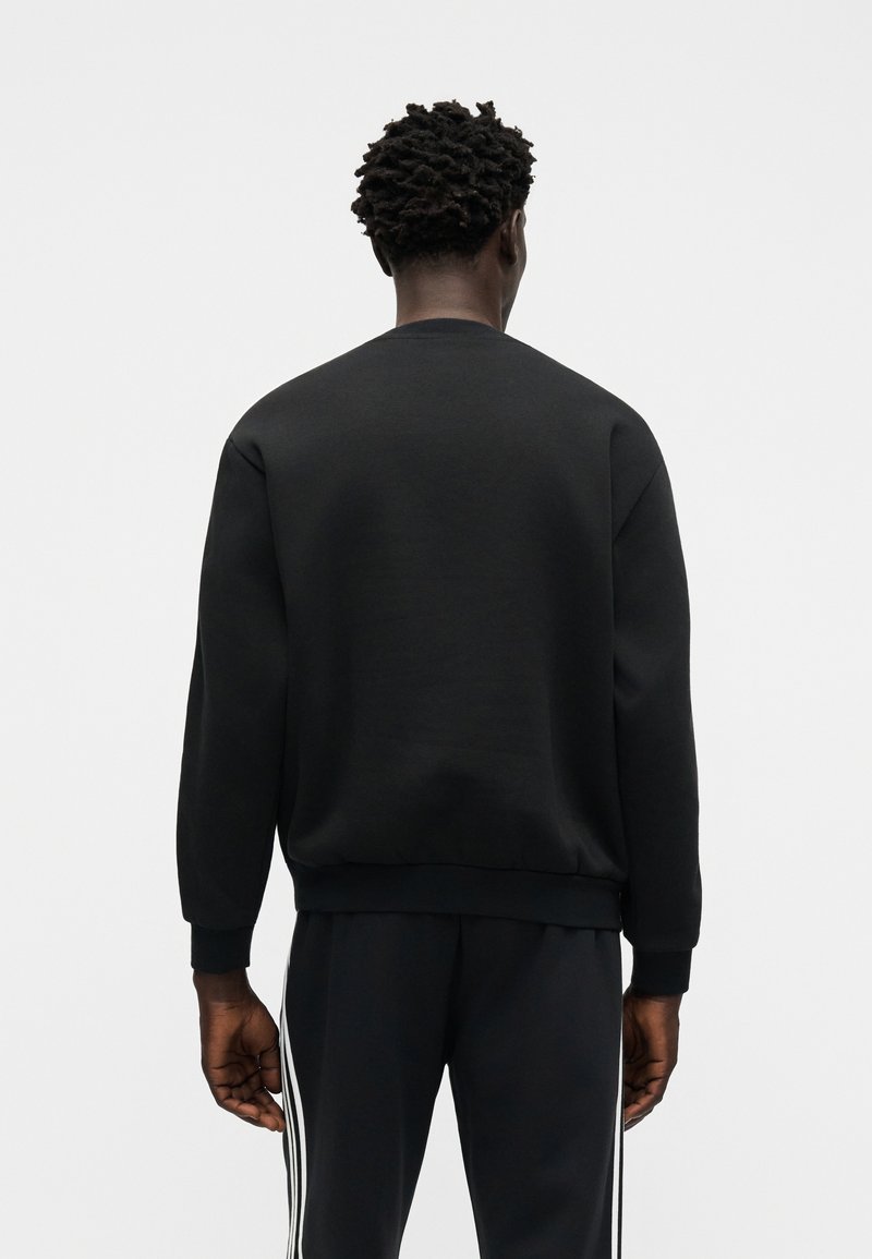 Man with short curly hair wearing a black sweatshirt and black pants with white stripes stands facing away against a plain background.