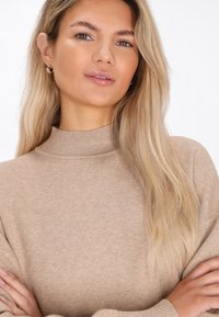Beige Rollkragenpullover mit weicher Textur und geripptem Kragen, mit langen Ärmeln und lässiger Passform, mit verschränkten Armen getragen.