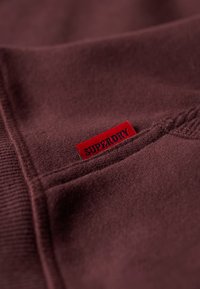 Tessuto in pile borgogna con una texture cucita, con una piccola etichetta rossa con la scritta "SUPERDRY" ben visibile.