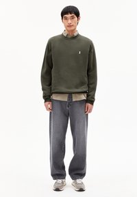 Olijfgroene sweatshirt met ronde hals en geborduurd wit logo, laag over een beige shirt, gecombineerd met losse grijze denim jeans en sneakers.