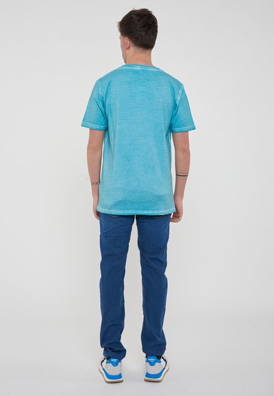 Camicia turchese a maniche corte con una finitura leggermente texture, abbinata a jeans blu. La camicia presenta cuciture a contrasto e un design con scollo rotondo.