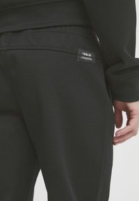 Jogging noir en tissu texturé, avec une étiquette logo sur la poche arrière et une taille lisse. Vue rapprochée du côté et de l'arrière.