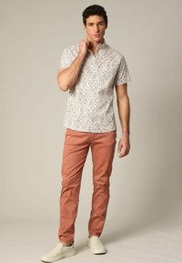 Chemise blanche à manches courtes avec un motif, associée à un pantalon corail clair et des chaussures à enfiler blanc cassé, modèle debout sur un fond neutre.