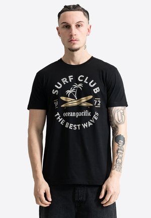 Muškarac s pletenicama i tetovažama na rukama nosi crnu Surf Club majicu s grafikom palmi i dasaka za surfanje.
