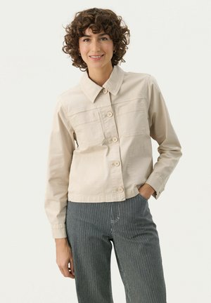 Beige kort jacka med knappknäppning fram, två bröstfickor och klassisk krage, i kombination med blå och vita randiga byxor.
