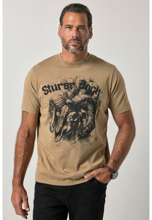 Homme adulte aux cheveux courts et à la barbe, portant un t-shirt beige avec un motif de crâne de bélier et un pantalon noir, debout avec une main dans la poche.