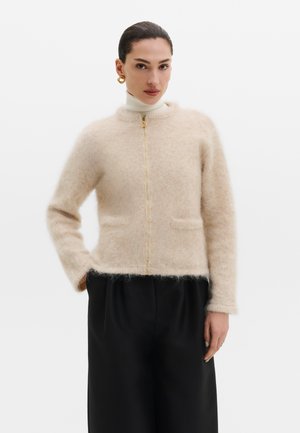 Femme portant un cardigan beige à fermeture éclair, duveteux, sur un col roulé blanc et un pantalon noir, avec des boucles d’oreilles anneaux dorés et les cheveux attachés.
