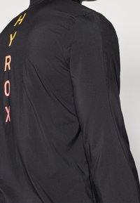 Chaqueta negra con una textura suave y ligera, con un logo de degradado vertical en amarillo y rosa en la parte posterior.