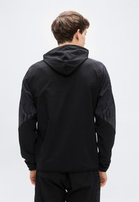 Μαύρο μπουφάν με κουκούλα, διακοσμημένο με μαύρες υφασμάτινες λεπτομέρειες που μοιάζουν με φτερά στα μανίκια. Σχεδιασμένο με εφαρμοστή γραμμή και λείο ύφασμα.