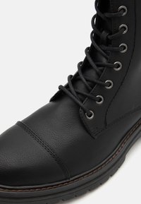Bota de cuero negro con cordones, punta redonda, ojales de metal plateado, superficie texturizada y costuras marrones en contraste a lo largo de la suela.