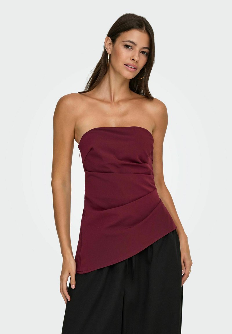 Burgundska brezramna majica z drapiranim oblikovanjem, ki se ponaša z gladko teksturo in asimetričnim robom. Združena z črnimi širokimi hlačami.