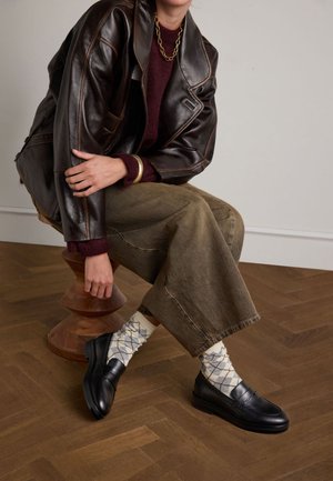 Zwarte leren instappers met een gladde textuur, gecombineerd met beige en blauwe argyle-geprinte sokken. Het model draagt olijfgroene wijde broek en een donker jack.