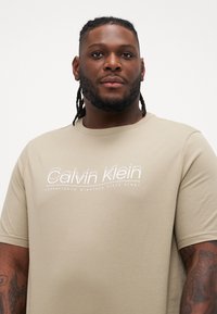 Bež pamukasta majica s kratkim rukavima i okruglim izrezom. Ima bijeli logo "Calvin Klein" otisnut sprijeda. Glatka tekstura.