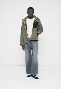 Giacca verde oliva con dettagli neri, cerniera e rifiniture arancioni. Indossata sopra una maglia bianca a coste, abbinata a jeans blu a gamba larga e scarpe da ginnastica nere.