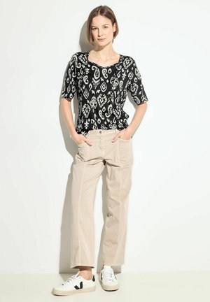 Zwart-wit gestreepte short-sleeve top, beige wijde broek en witte sneakers met zwarte accenten, staand tegen een lichte achtergrond.