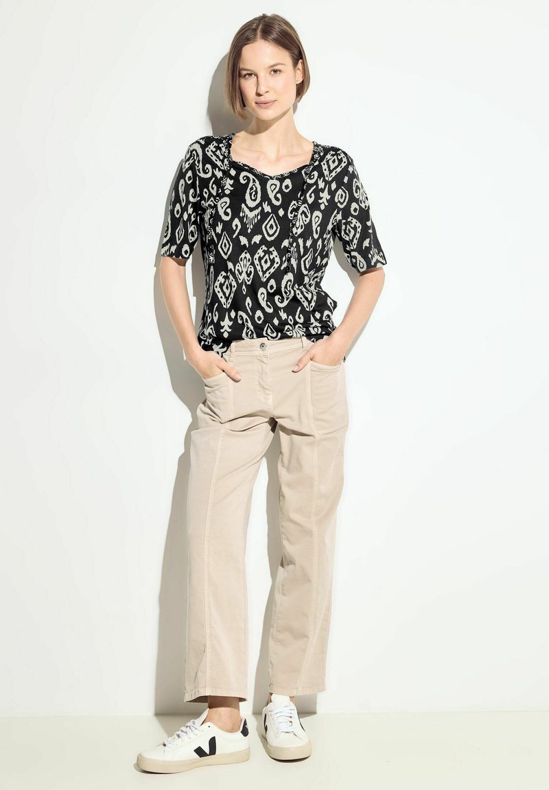 Top de manga corta con estampado en blanco y negro, pantalones de pierna ancha beige y zapatillas blancas con detalles en negro, de pie contra un fondo claro.